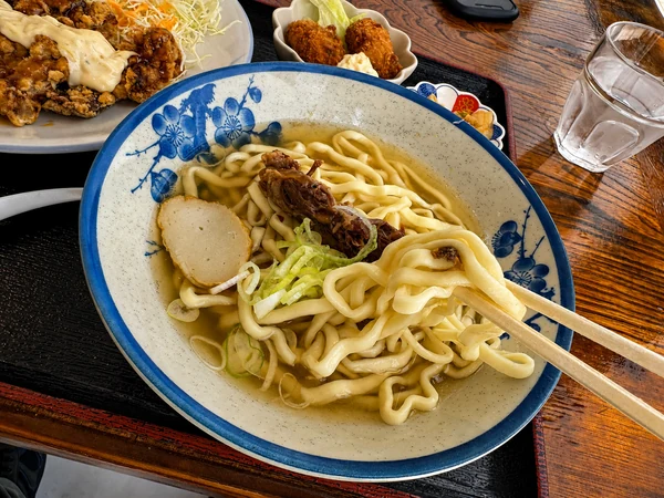 うるま市のうるまる食堂で提供された沖縄そばの麺を箸で持ち上げた様子。カツオ風味のあっさりスープと軟骨ソーキがのった一杯